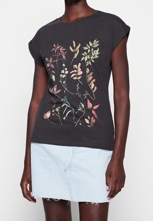 Camiseta de algodón negra con mangas cortas enrolladas, con un estampado botánico multicolor de hojas en la parte frontal. Combinada con una falda de mezclilla clara.