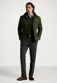 Man wearing dark green quilted jacket, blue shirt, dark sweater, gray trousers, and brown leather loafers, standing with hands in pockets.