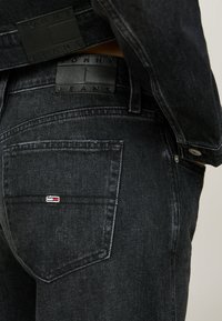 Vaqueros de mezclilla negros con una superficie texturizada. Presenta un bolsillo visible y un parche con el logotipo en la cinturilla. Colores de acento sutiles en las costuras.