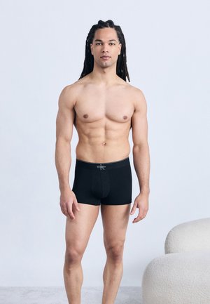 Vyras su ilgais drediškais, dėvintis juodus Calvin Klein trumpikius, stovintis prie šviesaus fono šalia baltos tekstūruotos kėdės.