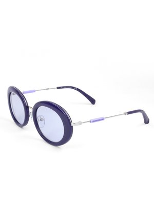 Occhiali da sole rotondi con montatura spessa in plastica viola scuro e lenti leggermente sfumate viola chiaro, aste sottili in metallo e logo "Calvin Klein Jeans" sui lati.