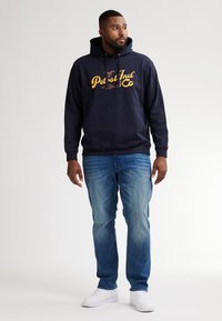 Felpa navy con logo giallo, tasca a marsupio e cordino regolabile. Abbinata a jeans azzurri chiari e sneakers bianche.