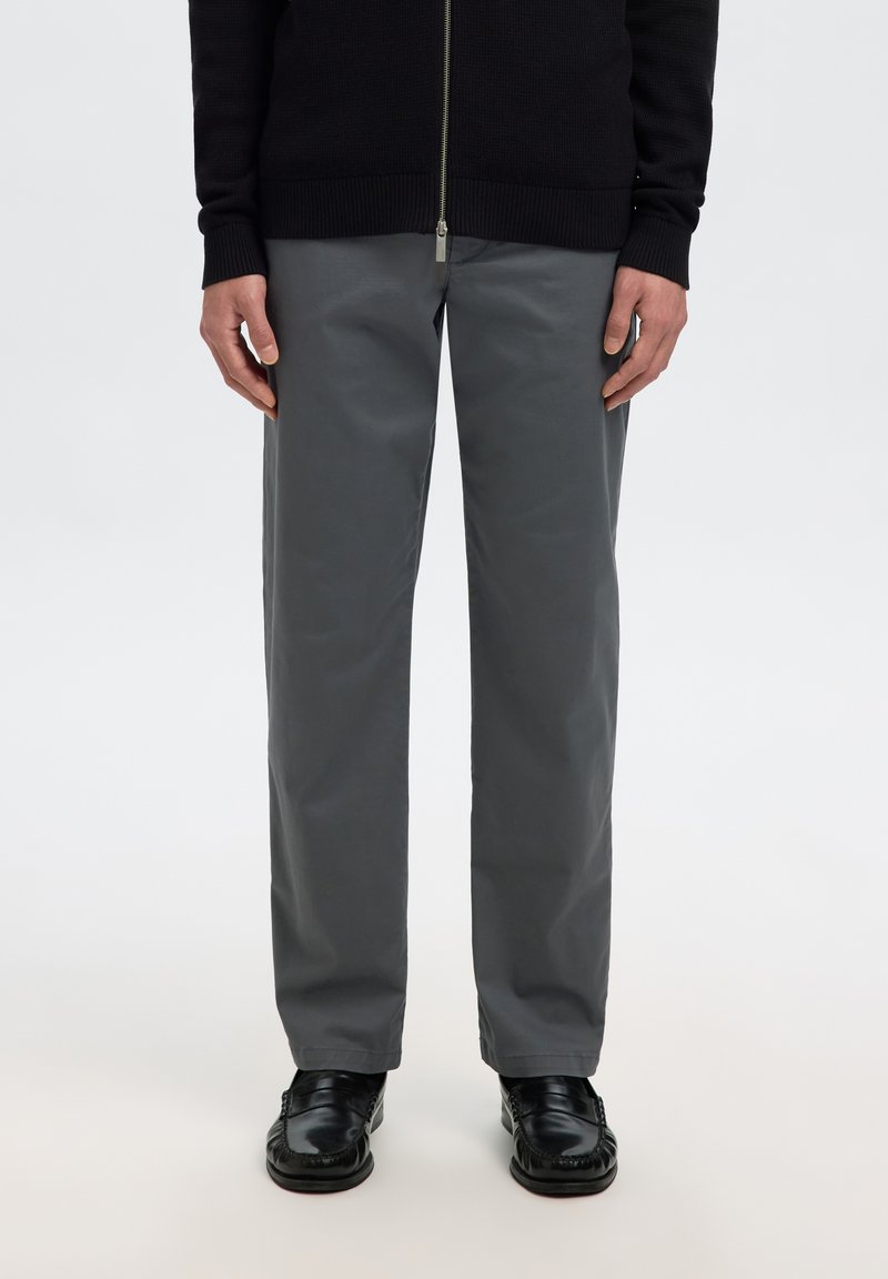 Personne portant un pantalon droit gris foncé, des mocassins en cuir noirs et un pull noir zippé, debout devant un fond uni.