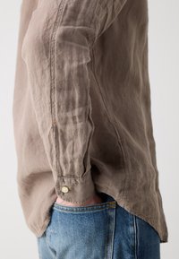 Bruine linnen overhemd met een gekreukt textuur en knoopmanchetten, gedragen over blauwe denim jeans. Het overhemd heeft een gebogen zoom.