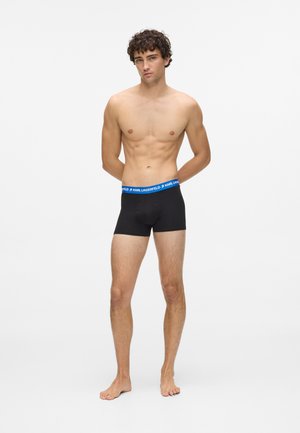 Giovane uomo in piedi a piedi nudi, senza maglietta, che indossa boxer neri con un elastico blu su cui è scritto "KARL LAGERFELD."