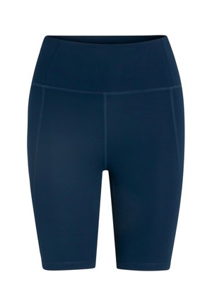 Χαμογελαστή γυναίκα με κόκκινα μαλλιά στέκεται φορώντας λευκό crop top, μαύρα ποδηλατικά σορτς με ψηλή μέση, λευκές κάλτσες και λευκά αθλητικά παπούτσια σε απλό φόντο.