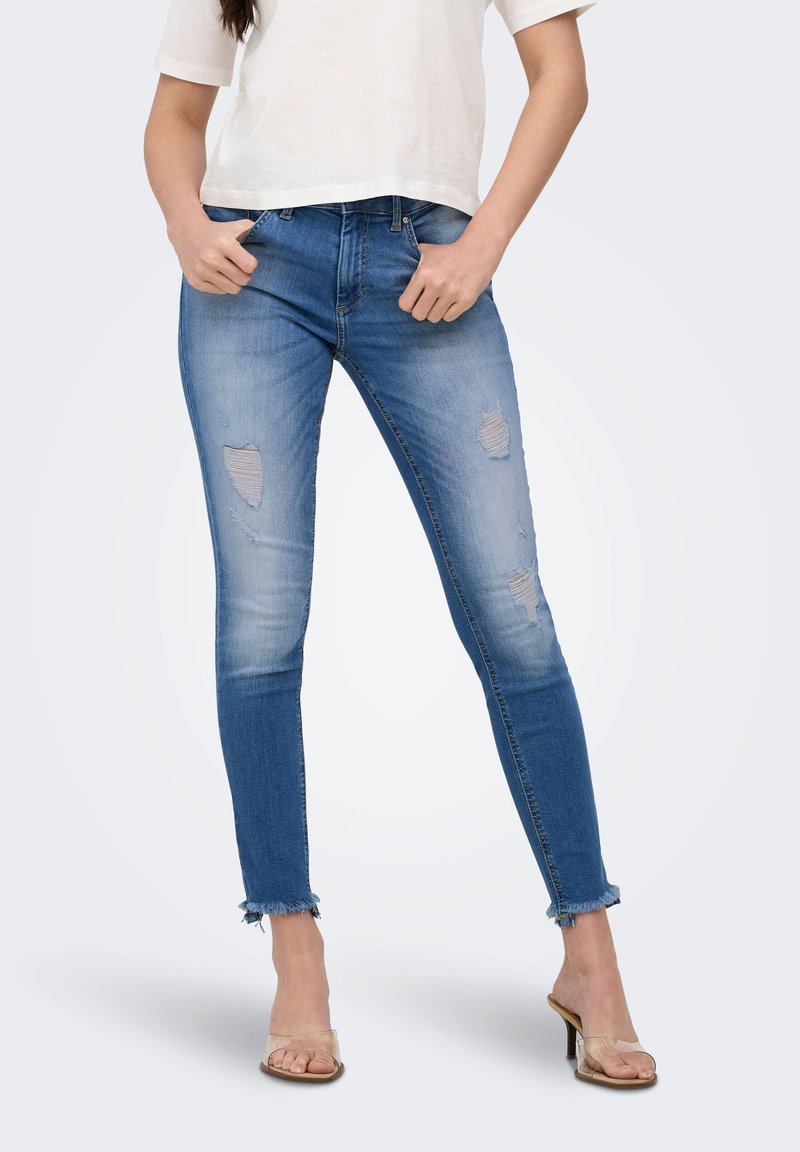 Femme portant un jean skinny bleu clair déchiré avec des ourlets effilochés, un t-shirt court blanc et des sandales à talons hauts transparentes, debout, les mains dans les poches.