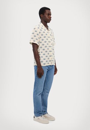 Short-sleeved, cream shirt with blue eye pattern, paired with light blue jeans and beige sneakers. Smooth fabric, relaxed fit.
