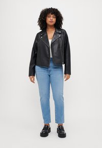 Schwarze Kunstleder-Bikerjacke mit silbernen Reißverschlüssen, getragen über einem weißen Oberteil. Kombiniert mit hellblauen Gerade geschnittenen Jeans und schwarzen Plateauschuhen.