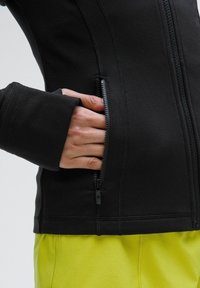 Veste zippée noire au design ajusté. Comprend une petite poche zippée à la taille et des ouvertures pour les pouces aux manches. Détail en tissu texturé.