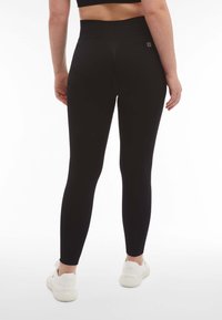 Leggings neri a vita alta con una superficie liscia, design aderente e un sottile logo sul cinturino, abbinati a sneakers chiare.