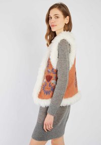 Brązowa kamizelka z białym puszystym wykończeniem, ozdobiona kolorową haftem kwiatowym. Noszona na szarej dzianinowej sukience. Prosty kształt i textured tkanina.