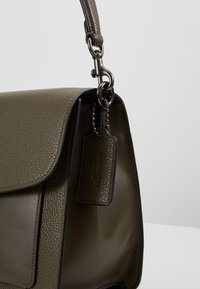 Sac à main en cuir vert olive avec textures granuleuse et lisse, doté d'une bandoulière amovible et d'une étiquette en cuir assortie sur une chaîne métallique.