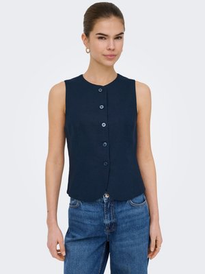 Femme portant un gilet sans manches bleu marine à boutons, associé à un jean bleu taille haute, debout devant un fond uni.