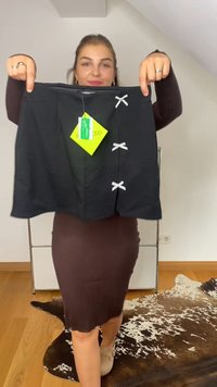 Kvinna klädd i en brun klänning håller upp en svart kjol med tre små vita rosetter och fästa gröna prislappar inomhus.