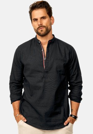 Mann mit Bart, der ein schwarzes, kragenloses Hemd mit rot-weißem Besatz, beige Hosen, eine Armbanduhr und ein Armband trägt, die Hände in den Taschen, neutraler Hintergrund.