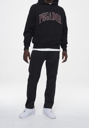 Muž ve černé mikině s nápisem "PEGADOR", černých kalhotách, s vrstvenou bílou košilí pod mikinou a bílých teniskách, stojící na bílém pozadí.