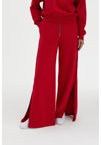 Rote weite Jogginghosen mit seitlichen Schlitz, aus weichem Stoff. Mit einer Reißverschlusstasche vorne und kombiniert mit weißen Sneakers.