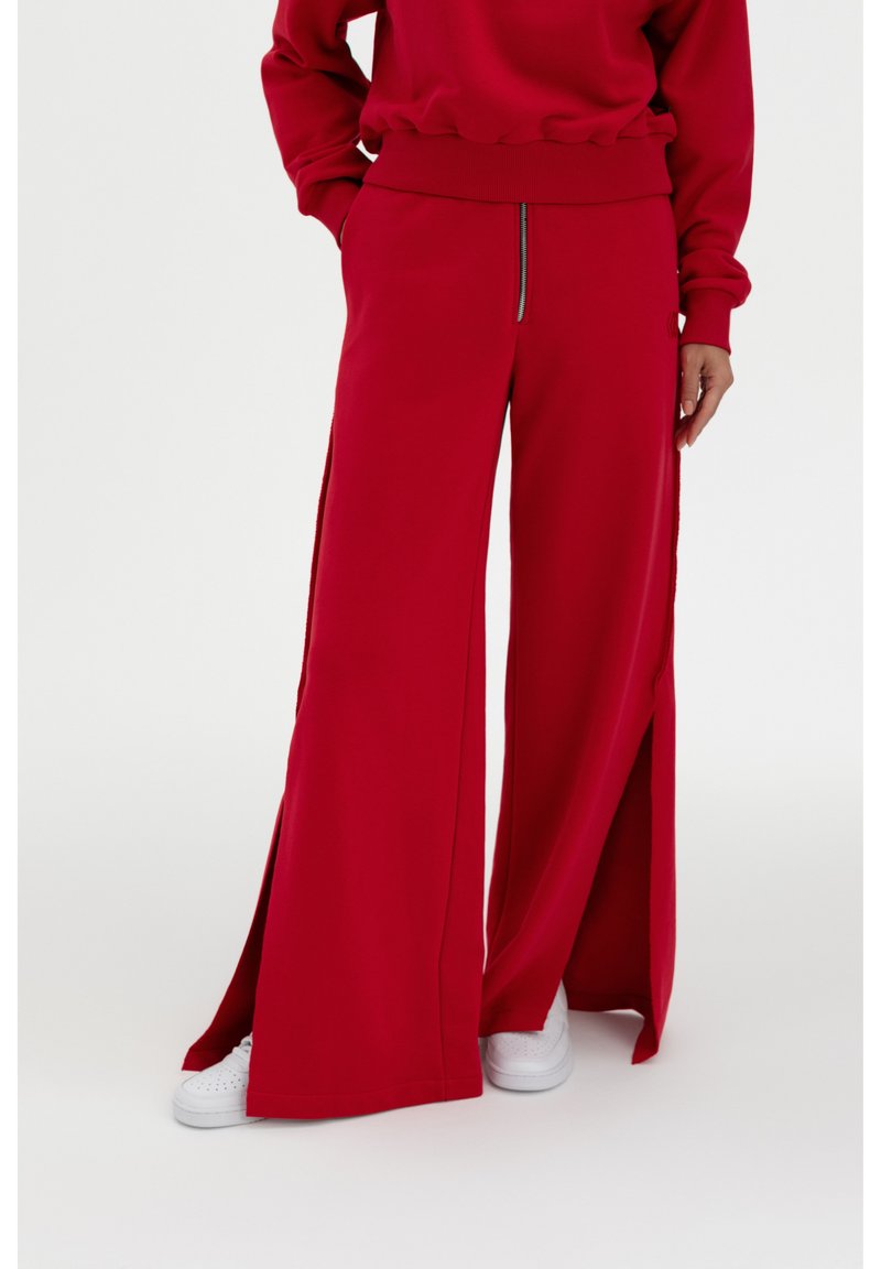 Rote weite Jogginghosen mit seitlichen Schlitz, aus weichem Stoff. Mit einer Reißverschlusstasche vorne und kombiniert mit weißen Sneakers.