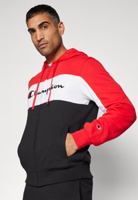 Sudadera con cremallera en rojo, blanco y negro, hecha de una mezcla de algodón; presenta un logo en la parte frontal, bolsillos tipo canguro y puños y dobladillo acanalados.