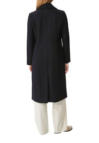 Manteau noir long avec col, tissu texturé et coupe droite, porté avec un pantalon crème, vue arrière.