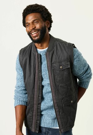 Man with curly hair and beard smiling, wearing a blue sweater and a black sleeveless quilted vest with pockets.