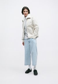 Giovane uomo che indossa una giacca di shearling color crema, una polo grigia chiara, jeans azzurri corti, calzini bianchi e mocassini neri, in piedi contro uno sfondo bianco.