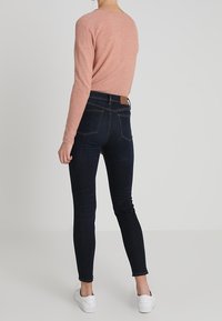 Růžový top s dlouhým rukávem, přiléhavý design, spojený s tmavě modrými skinny džíny a bílými teniskami. Jednoduchá, texturovaná látka s minimálními detaily.