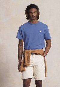 Young man with dreadlocks wearing a blue t-shirt, beige shorts, a brown sweater tied around waist, and a wristwatch, standing against a neutral background.