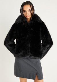 Jaqueta de pelo sintético preta com um colarinho largo e mangas longas, apresentando uma textura felpuda. Usada sobre uma saia cinza com uma fenda lateral.