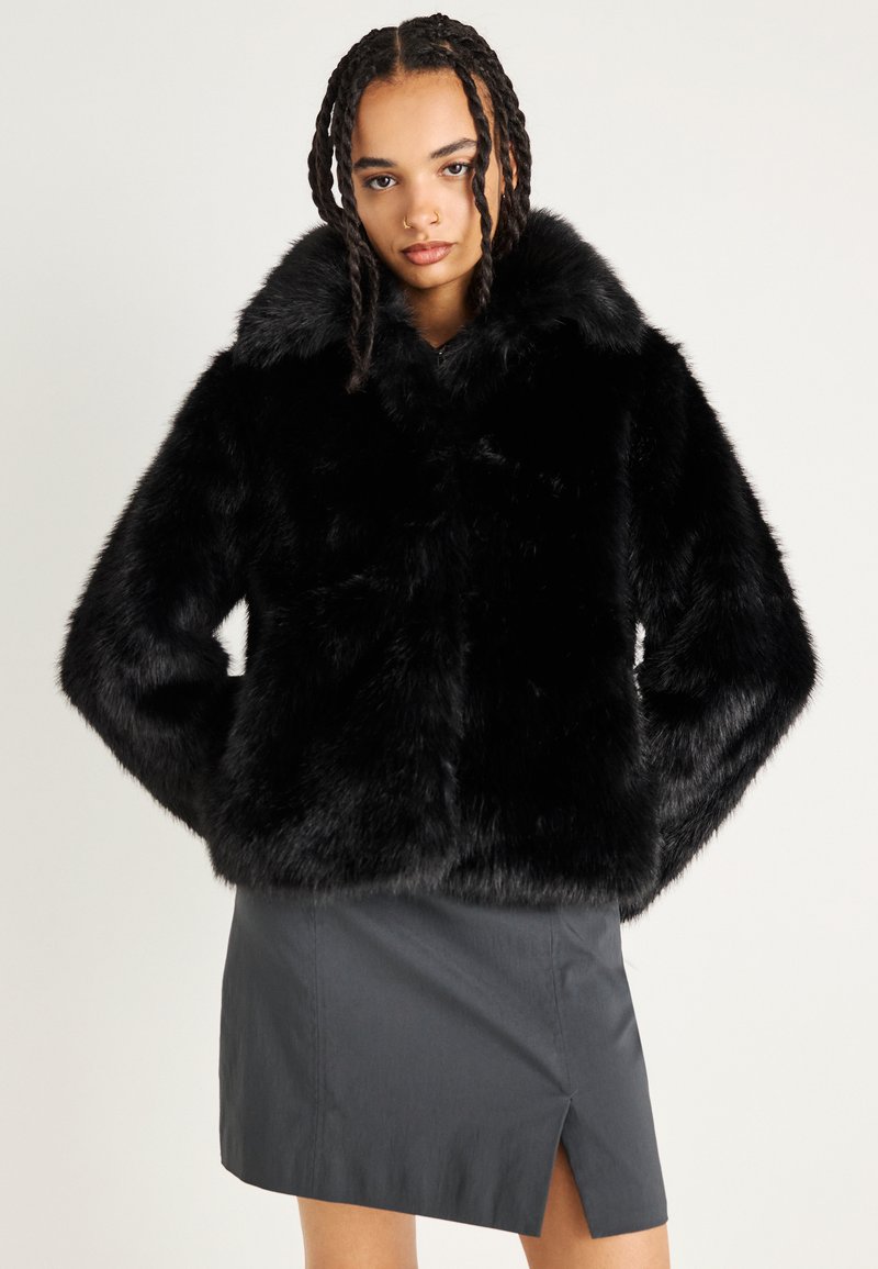 Melnas mākslīgās kažokādas jakas ar plašu apkakli un garām piedurknēm, kas iezīmējas ar greznu tekstūru. Valkāta virs pelēka svārku ar sānu šķirumu.