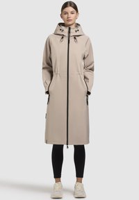 Parka impermeabil bej, până la genunchi, cu fermoar negru, glugă cu șnur, buzunare laterale și manșete elastice. Textură fină, design minimal.