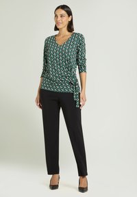 Groen top met wikkelpatroon, een V-hals, lange mouwen en een zijstrik. Gecombineerd met rechte zwarte broek en zwarte hakken.