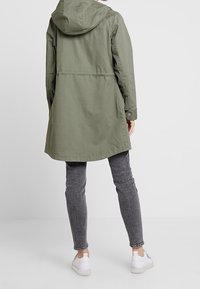 Olivově zelená bunda s kapucí, volným střihem, dlouhými rukávy a střední délkou. Má knoflíkové detaily a kapsy po stranách.