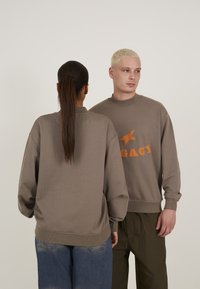 Hanorace maro supradimensionate cu mansete ribbate, având text grafic portocaliu pe față. Material texturat; purtate cu blugi largi albaștri și pantaloni verzi.