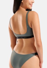 Conjunto de bañador verde con textura suave y brillante; incluye un bralette de escote bajo con banda negra con logo y braguitas de corte alto a juego.