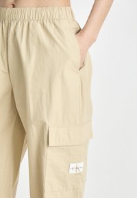 Beige cargobroek met een elastische tailleband, zijzak en logo-opdruk op de bovenbeen. Het materiaal lijkt glad en licht van gewicht.