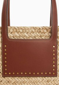 Woven straw handbag z prostokątną brązową skórzaną łatką, ozdobioną złotymi ćwiekami wzdłuż krawędzi oraz dwiema skórzanymi uchwytami przeszytymi nitkami.