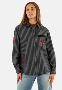 Donna che indossa una camicia nera a righe con bottoni, con toppe e ricami rossi, abbinata a jeans azzurri, in piedi davanti a uno sfondo bianco.