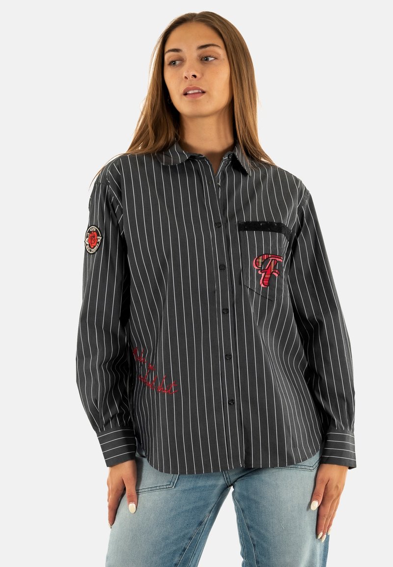 Donna che indossa una camicia nera a righe con bottoni, con toppe e ricami rossi, abbinata a jeans azzurri, in piedi davanti a uno sfondo bianco.