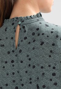 Blouse verte avec un col à volants, comportant une ouverture en forme de goutte d'eau dans le dos et un motif de taches noires sur le tissu.