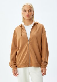 Sudadera con cremallera marrón de textura suave, capucha y puños acanalados. Combinada con una camisa blanca, sobre un fondo liso.