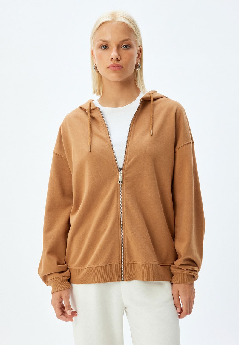 Sudadera con cremallera marrón de textura suave, capucha y puños acanalados. Combinada con una camisa blanca, sobre un fondo liso.