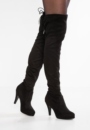 Kobieta w czarnych zamszowych wysokich kozakach za kolano na obcasie, z ostrymi noskami i sznurkiem do zawiązywania u góry, stojąca na białej podłodze.