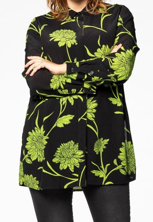 Person iført en sort skjorte med lyst grønt blomsterprint, arme krydset over brystet, og sorte bukser mod en hvid baggrund.