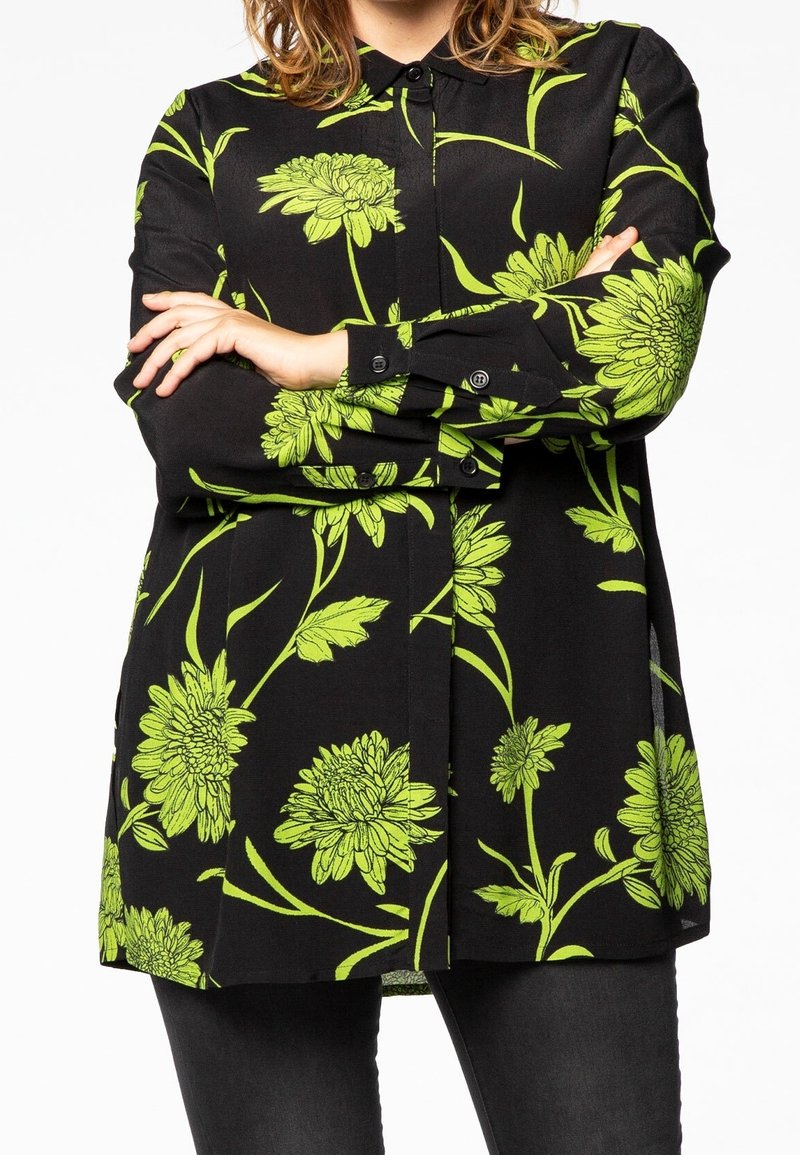 Personne vêtue d'une chemise noire à imprimé floral vert vif, bras croisés sur la poitrine, et pantalon noir sur fond blanc.