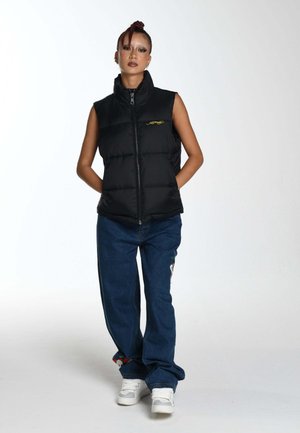 Chaleco acolchado negro con cuello alto, cierre completo y bordado de logo amarillo. Combinado con pantalones de mezclilla azul y zapatillas blancas.
