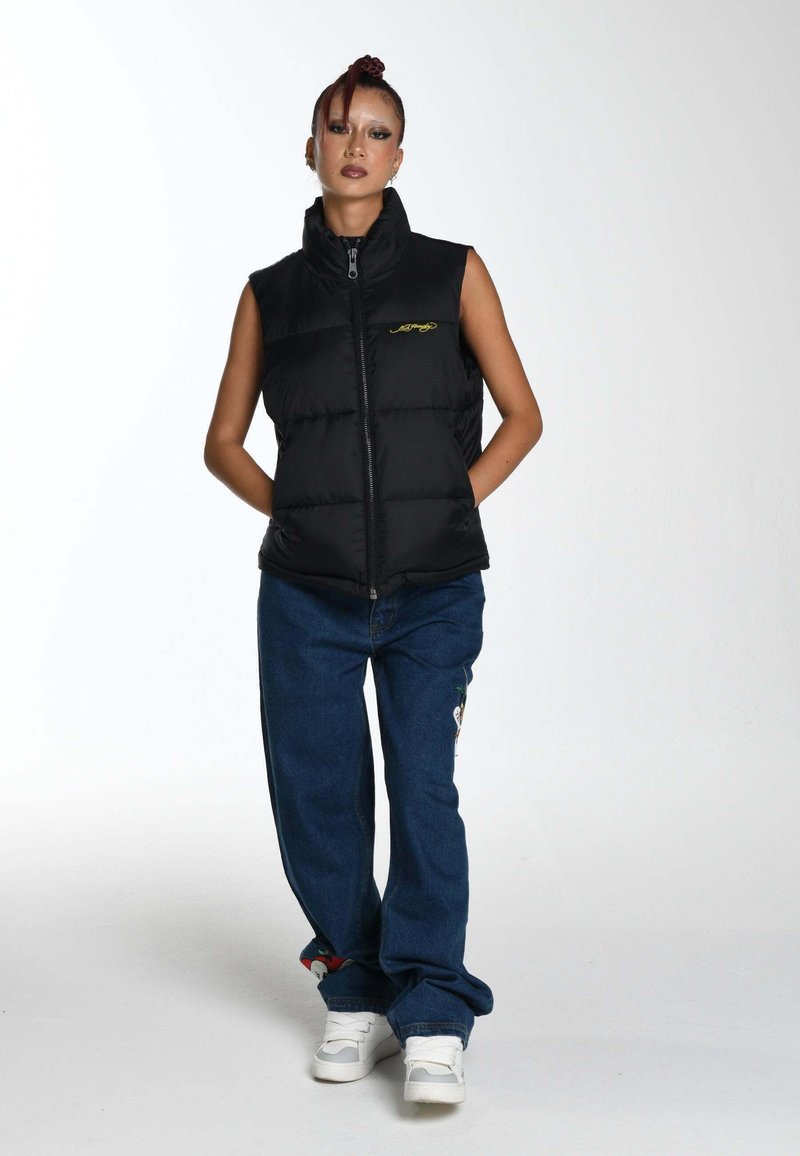 Chaleco acolchado negro con cuello alto, cierre completo y bordado de logo amarillo. Combinado con pantalones de mezclilla azul y zapatillas blancas.