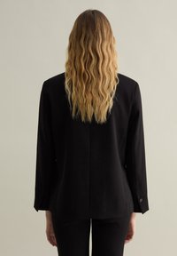 Blazer noir à manches longues, design structuré, accents de boutons sur les poignets, texture lisse et détail d'ouverture à l'arrière.