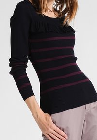 Navy ribbed knit long-sleeve sweater with purple horizontal stripes and ruffle detail at the neckline. Fitted design with a rounded hem.